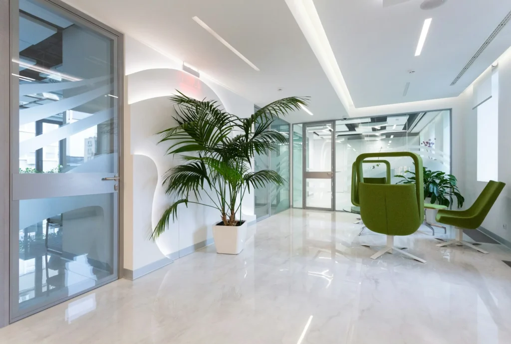 A bright, modern office lobby with a potted palm, glass walls, and green accent chairs