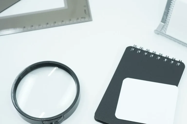 A magnifying glass and a closed spiral notebook on a white desk